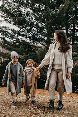 Mother and two children, all wearing coats and scarves, walking hand in hand through a forested area on a cloudy day