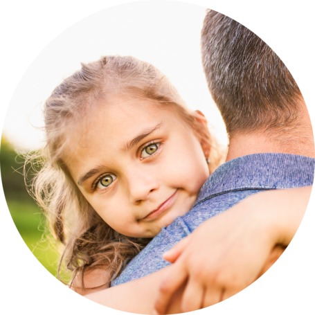 Young girl with a ponytail being hugged by an adult, looking over their shoulder with a gentle expression, in an outdoor setting.