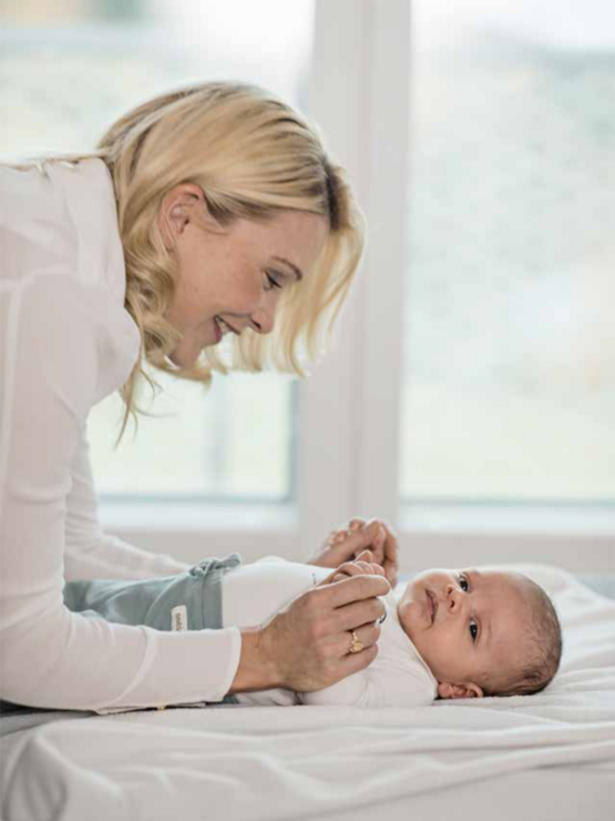 Smiling woman leaning over a changing table, looking at a baby lying down and dressed in a white onesie, with a window in the background.